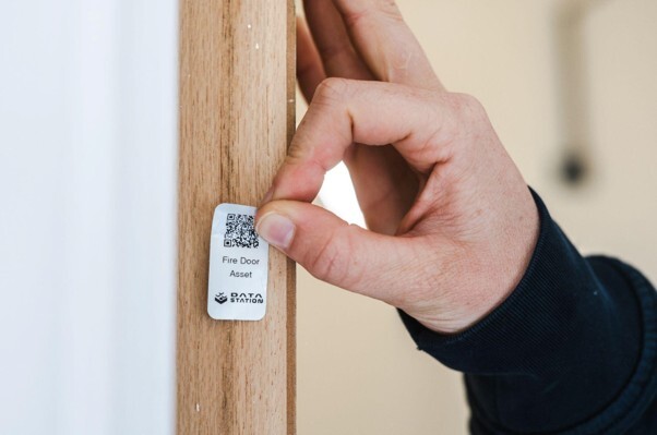 Fire door compliance checklist inspection with an asset label being checked on a wooden fire door.
